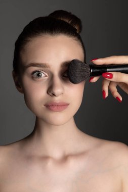 Gri stüdyo arka planında izole edilmiş makyaj fırçası olan beyaz bir kızın yakın plan portresi. Dikey resim