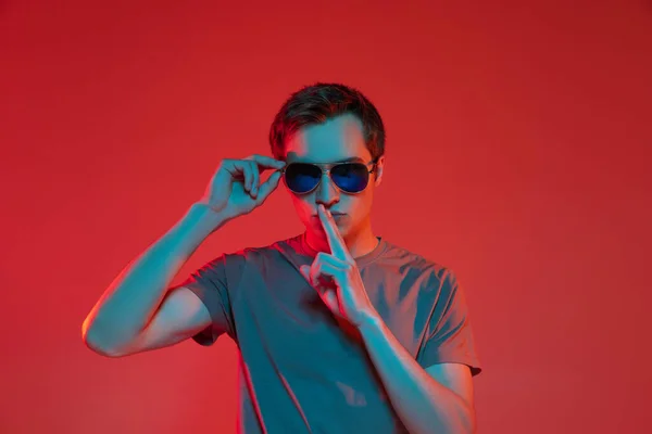 Jeden młody, przystojny biały facet w okularach przeciwsłonecznych, odizolowany na czerwonym neonowym tle. Pojęcie ludzkich emocji, wyraz twarzy. — Zdjęcie stockowe