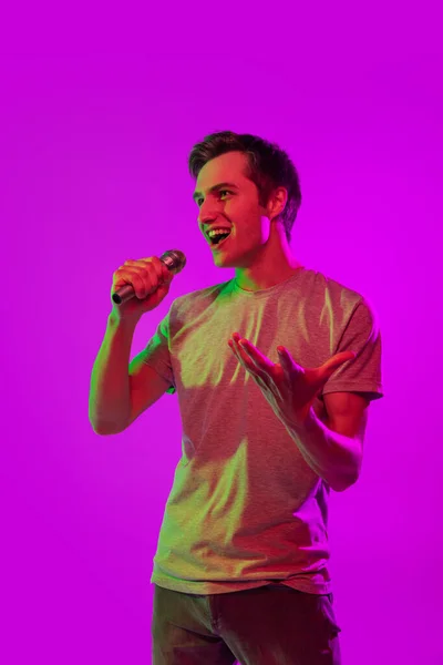 Un giovane bel ragazzo sorridente con microfono isolato su sfondo rosa al neon. Concetto di espressione facciale ed emozioni umane — Foto Stock