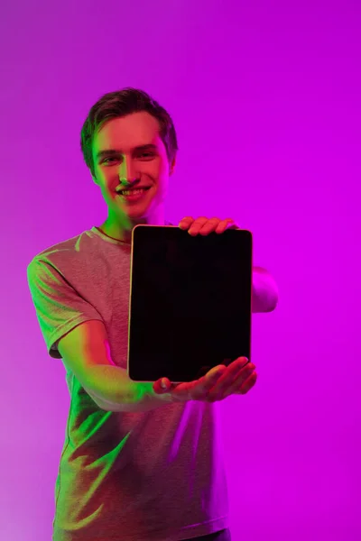 Studio image of young caucasian man with digital tablet on pink background in neon light. Concept of emotions, youth, sales — Stock Photo, Image