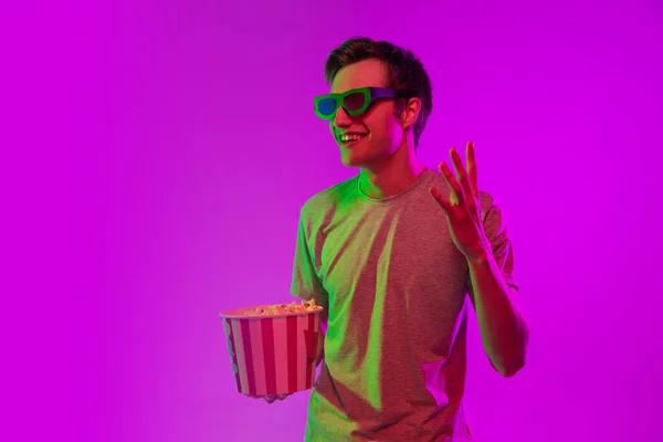 Βλέποντας κινηματογράφο. Νεαρός καυκάσιος άνδρας με ποπ κορν σε 3d ποτήρια απομονωμένος σε ροζ φόντο στούντιο σε νέον φως. — Φωτογραφία Αρχείου