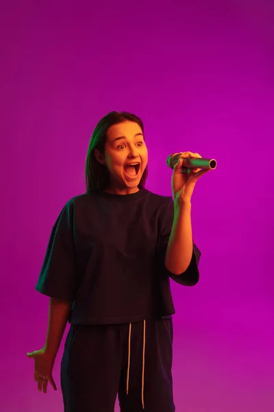 Retrato vertical de una chica caucásica bastante joven cantando emocionalmente en el micrófono sobre fondo rosa en luces de neón —  Fotos de Stock