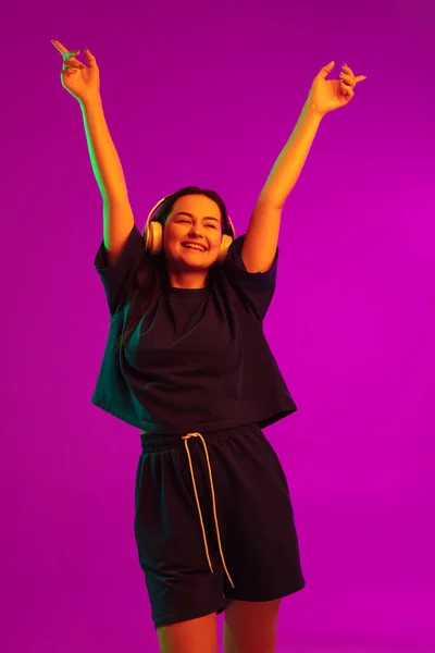 Studioaufnahme eines jungen hübschen kaukasischen Mädchens mit Kopfhörern, das isoliert auf rosa Hintergrund im Neonlicht zur Musik tanzt — Stockfoto