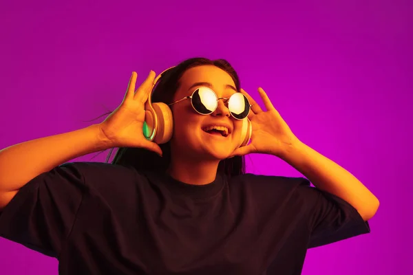 Mi piace la musica. Giovane bella ragazza caucasica in cuffia e occhiali da sole luminosi isolati su sfondo rosa in luci al neon — Foto Stock