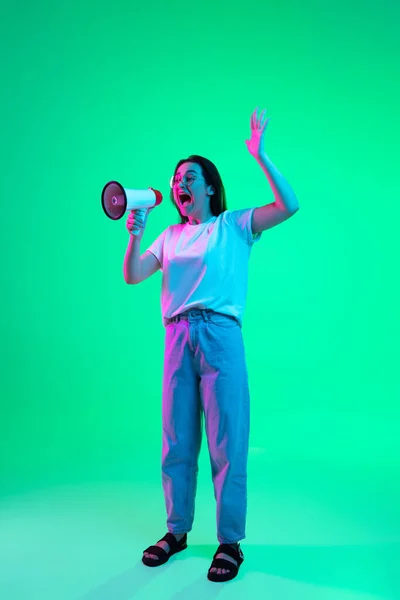 Retrato de larga duración de la joven hermosa chica caucásica en ropa casual con megáfono aislado sobre fondo verde en luz de neón. — Foto de Stock