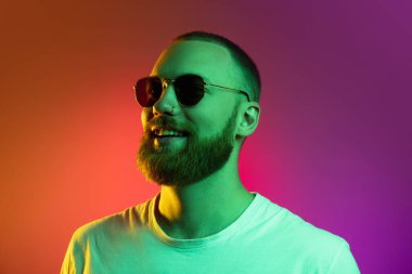 Güneş gözlüklü yakışıklı sakallı genç bir adam. Neon ışıklarıyla izole edilmiş turuncu bir arka planda..