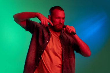 Mikrofonlu beyaz sakallı genç bir adamın stüdyo görüntüsü. Neon ışıklarıyla yeşil arkaplanda izole edilmiş..