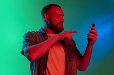 Akıllı telefonlu beyaz sakallı genç bir adamın stüdyo görüntüsü. Neon ışıklarıyla yeşil arka planda izole edilmiş..