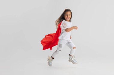 Küçük süper kahraman. Kırmızı pelerinli beyaz kız beyaz stüdyo arka planında izole edilmiş. Yan görünüm