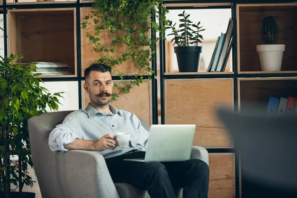 One serious young businessman sitting in comfortable armchair with laptop and cup of coffee. Front view — Fotografia de Stock