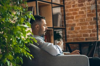Working, drinking coffee. Image of one young businessman sitting in comfortable armchair with laptop and cup of coffee.