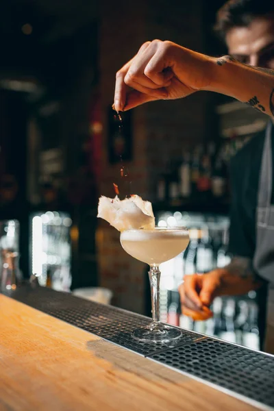 One young professional bartender decorating alcohol cocktail with flakes or dried flowers standing at bar counter — Foto de Stock