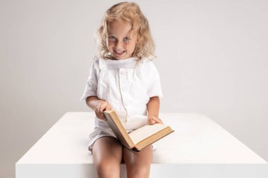 Beyaz stüdyo arka planında izole edilmiş büyük bir kutunun üzerinde oturan beyaz bir kız kitap okuyor..