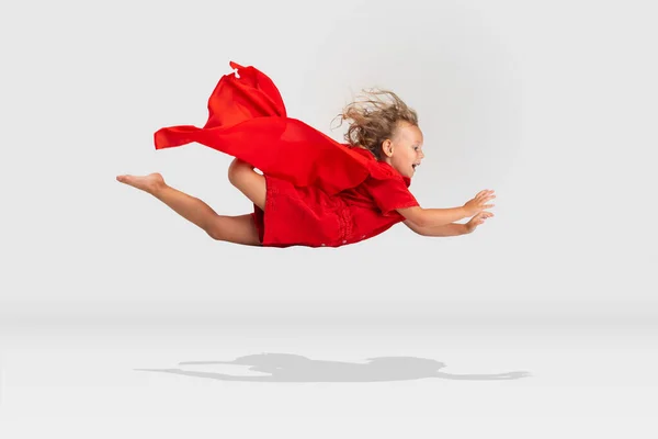 Jonge superheld. Schattig klein lachend meisje in rode cape vliegend geïsoleerd over witte studio achtergrond. Zijaanzicht — Stockfoto