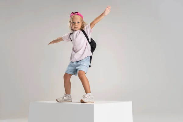 En liten förskola kaukasiska flicka i casual kläder med ryggsäck står på stor låda isolerad över vit studio bakgrund. — Stockfoto