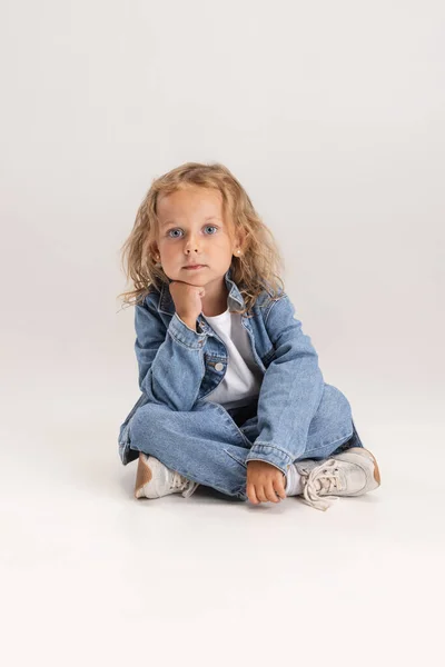 Porträtt av liten förskola kaukasiska flicka i denim kläder sitter på golvet isolerad över vit studio bakgrund. — Stockfoto