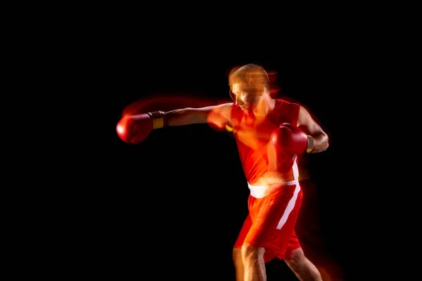 En professionell boxare i röda shorts och handskar träning, tränar över svart bakgrund i blandade ljus — Stockfoto