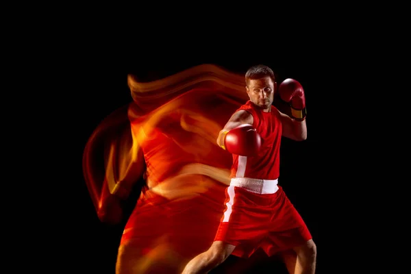 Joven boxeador profesional masculino en pantalones cortos rojos y guantes de entrenamiento, ejercitando sobre fondo negro en luces mixtas — Foto de Stock