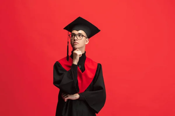 Young caucasian guy, student, graduate in mantle and mortarboard isolated over red studio background. — Zdjęcie stockowe