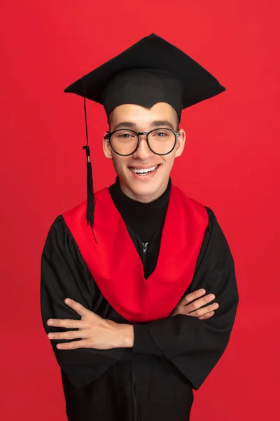Studio shot of young smiling, cheerful student, graduate in mortarboard isolated over red studio background. — Stock Fotó