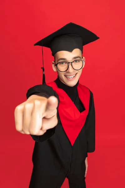 Studio shot of young smiling, cheerful student, graduate in mortarboard isolated over red studio background. — Stockfoto