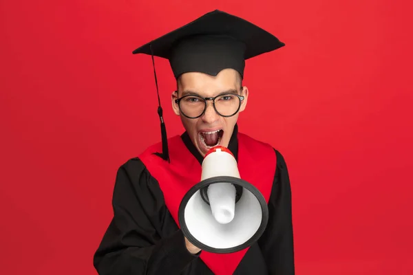 Portrait of young student, graduate in mantle with megaphone isolated over red studio background. — Stok fotoğraf