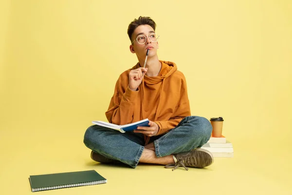 One young caucasian guy, student sitting on floor with notepad and pen isolated over yellow studio background. — Stock Fotó