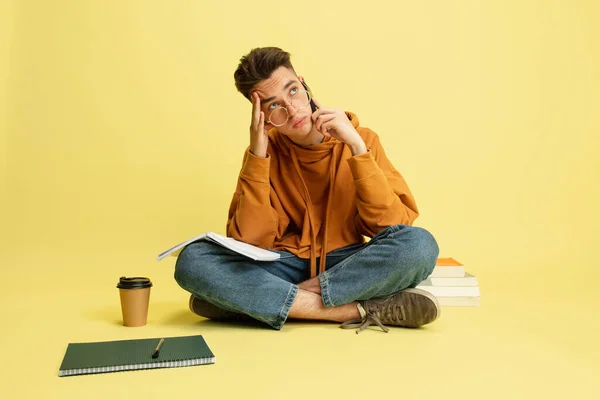 One young caucasian guy, student sitting on floor and doing homework isolated over yellow studio background. — Fotografia de Stock
