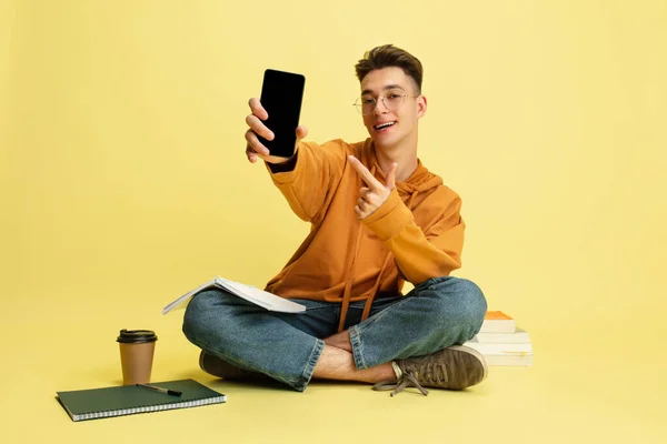 一个年轻的高加索人，坐在地板上看书，手持智能手机，隔离在黄色的工作室背景下. — 图库照片