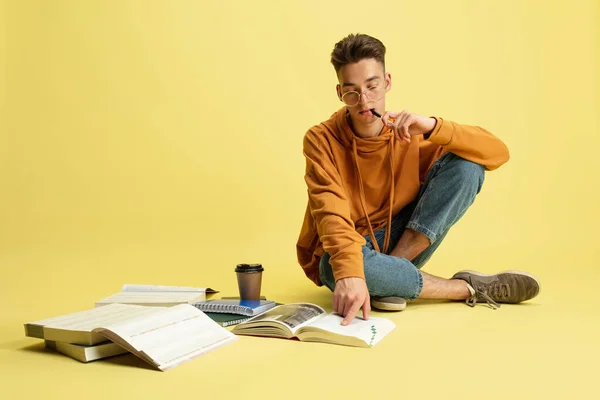 Portrait of young caucasian guy, student sitting on floor with books, notepad isolated over yellow studio background. — Stock Fotó