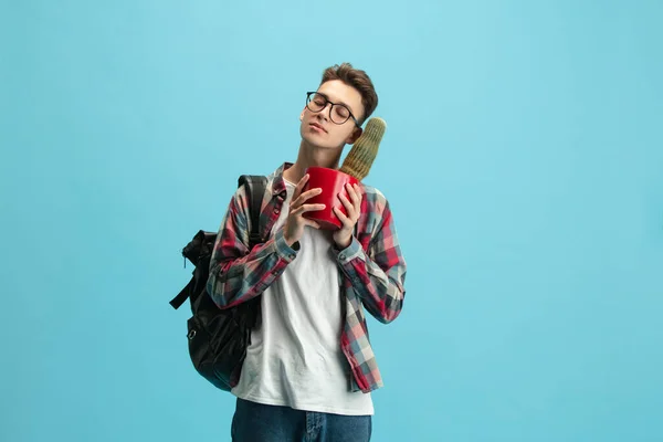 One young caucasian guy, student with backpack and cactus in a pot isolated over light blue studio background. —  Fotos de Stock