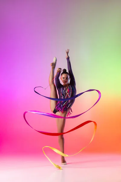 Verticaal portret van jonge vrouwelijke ritmische gymnaste oefenen met gekleurd lint geïsoleerd op heldere neon achtergrond. — Stockfoto