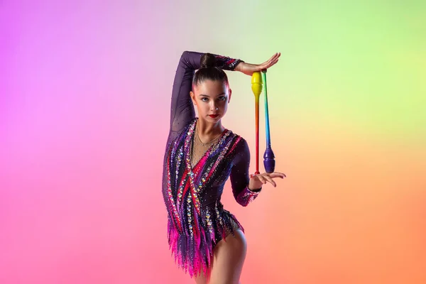 Una gimnasta rítmica femenina con clubes posando aislados sobre fondo de neón multicolor. Retrato de media longitud — Foto de Stock