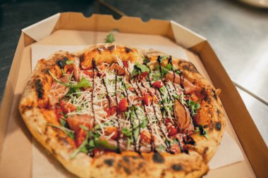 Etli, jambonlu, yeşil ve domatesli fırında pizza. Karton kutuya sos serpiştirilmiş..