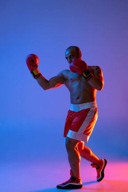 Kırmızı şortlu profesyonel boksörün dikey portresi neon ışıkta mor arka planda egzersiz yapıyor. Tam uzunluk