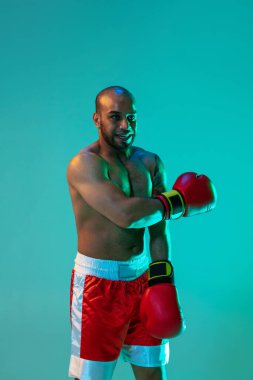 Dikey görüntü. Kırmızı şortlu ve eldivenli bir profesyonel boksör mavi yeşil arka planda neon ışıkta poz veriyor..