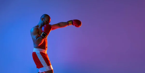En professionell boxare i röda shorts träning, tränar över lila bakgrund i neonljus. Sidovy — Stockfoto
