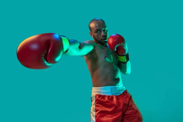 Silné pěsti. Jeden profesionální boxer v červených trenýrkách trénink, cvičení izolované přes modré zelené pozadí v neonovém světle. — Stock fotografie