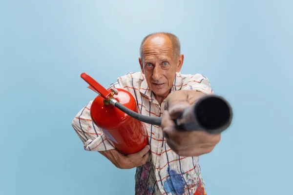 Peintre gris mature ou senior avec extincteur debout devant la caméra. Homme drôle dans une chemise teintée de peinture folie autour isolé sur bleu — Photo