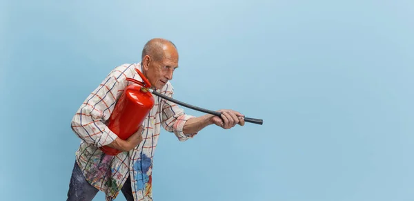 Pintor gris maduro o senior con extintor de incendios delante de la cámara. Hombre gracioso en una camisa manchada de pintura tonteando alrededor aislado en azul — Foto de Stock