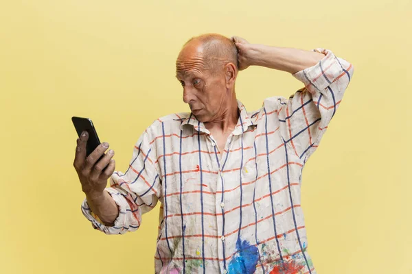 Mature or senior grey painter with a phone standing in front of camera. Funny man in a paint-stained shirt fooling around isolated on yellow — Stock Photo, Image