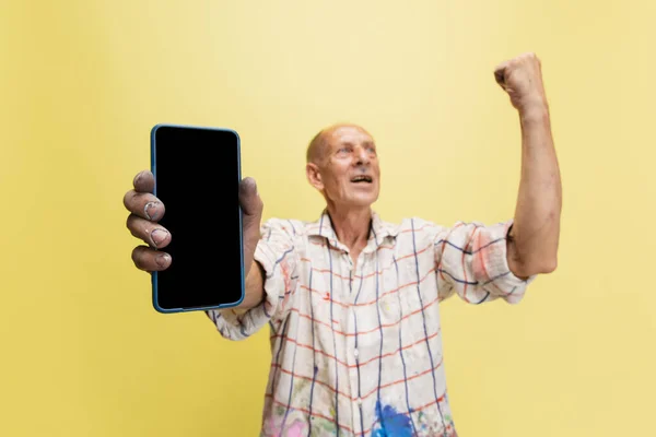 Peintre gris mature ou senior avec un téléphone debout devant la caméra. Homme drôle dans une chemise teintée de peinture s'amuser autour isolé sur jaune — Photo