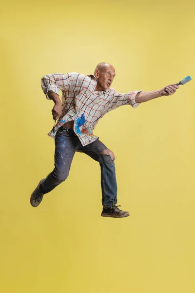 Dojrzały lub starszy szary malarz ze szczotką w skoku przed kamerą. Zabawny facet w poplamionej farbą koszuli zabawia się na żółto. — Zdjęcie stockowe