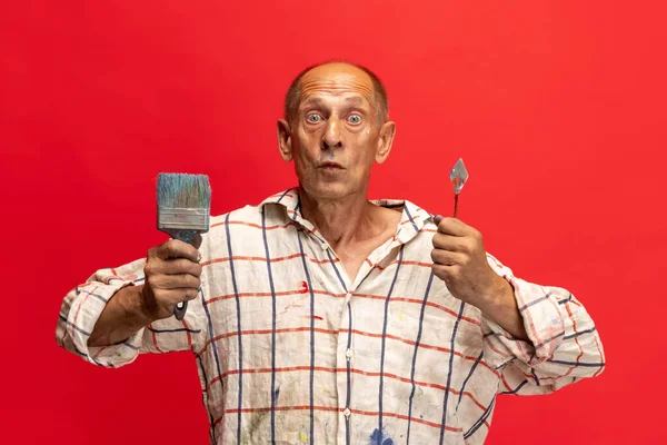 Dojrzały lub starszy szary malarz ze szczotką stojącą przed kamerą. Zabawny facet w poplamionej farbą koszuli zabawia się na czerwono — Zdjęcie stockowe