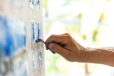 Olgun ya da yaşlı gri boyacı elinde fırçayla kameranın önünde duruyor. Stüdyoda ya da evde çalışan boyalı gömlekli bir adam.