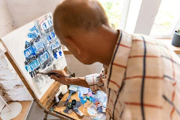 Pintor gris maduro o senior con un pincel delante de la cámara. Hombre serio con una camisa manchada de pintura que trabaja en el estudio o en casa — Foto de Stock