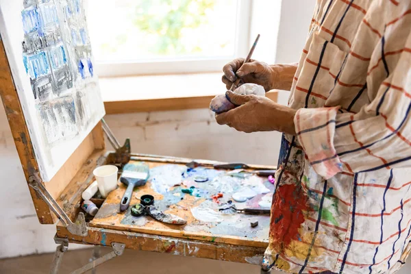 Pintor cinza maduro ou sênior com um pincel em pé na frente da câmera. homem com uma camisa manchada de tinta trabalhando em estúdio ou em casa — Fotografia de Stock