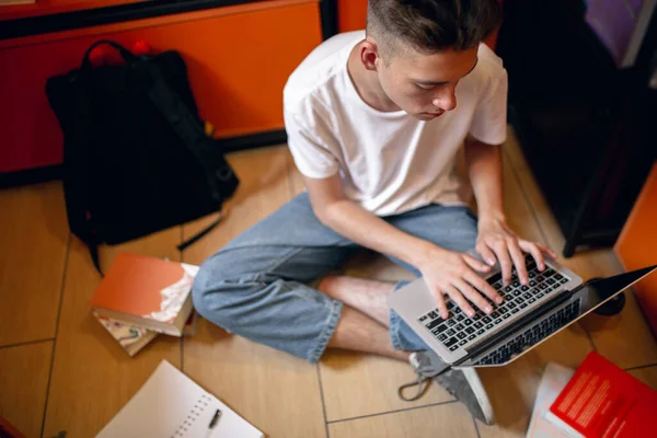 Un giovane ragazzo caucasico, studente seduto al piano con computer portatile su sfondo casa o biblioteca. — Foto Stock