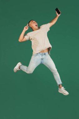 Koyu yeşil stüdyo arka planında izole edilmiş akıllı telefonlu yakışıklı Asyalı bir adamın portresi..