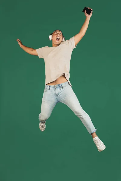 Portrait of young handsome asian man in headphones jumping with phone isolated over dark green studio background. — ストック写真